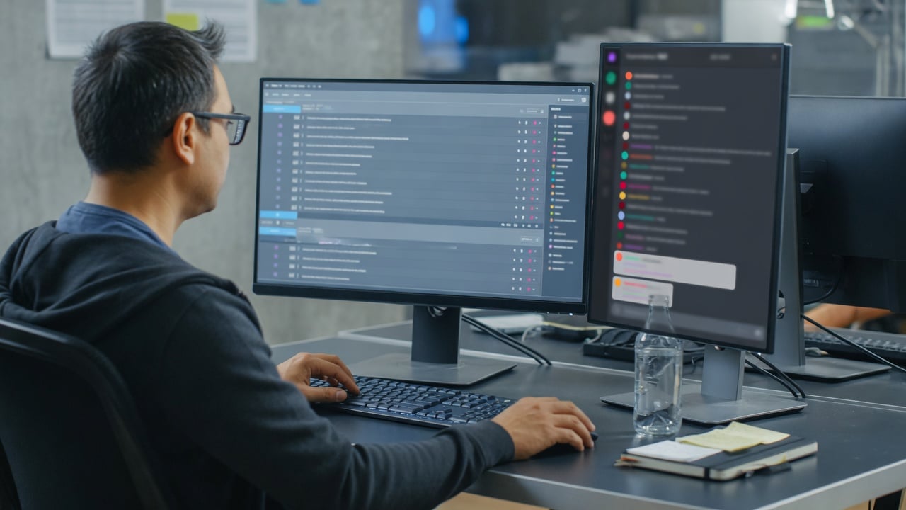 Administrator reviewing a forum thread list beside a muted chat sidebar on a dual-monitor desk