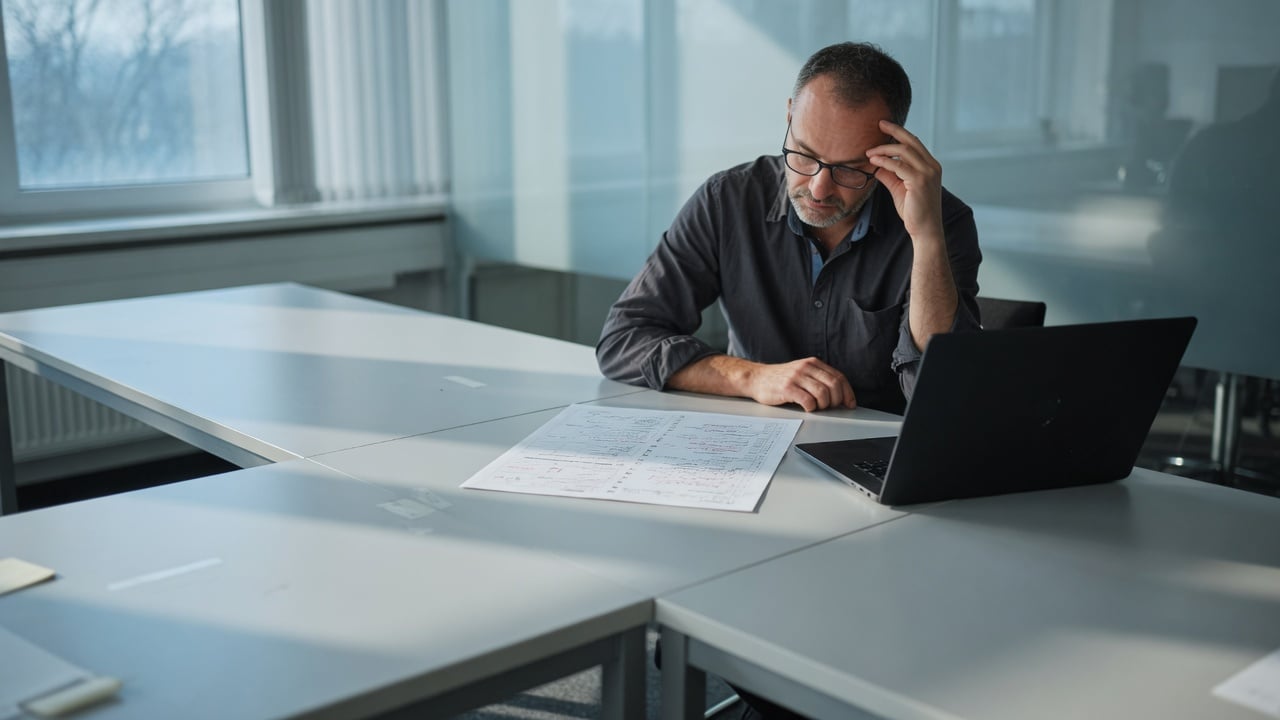 Administrator at a desk with a printed decision checklist beside a laptop showing a muted forum admin screen